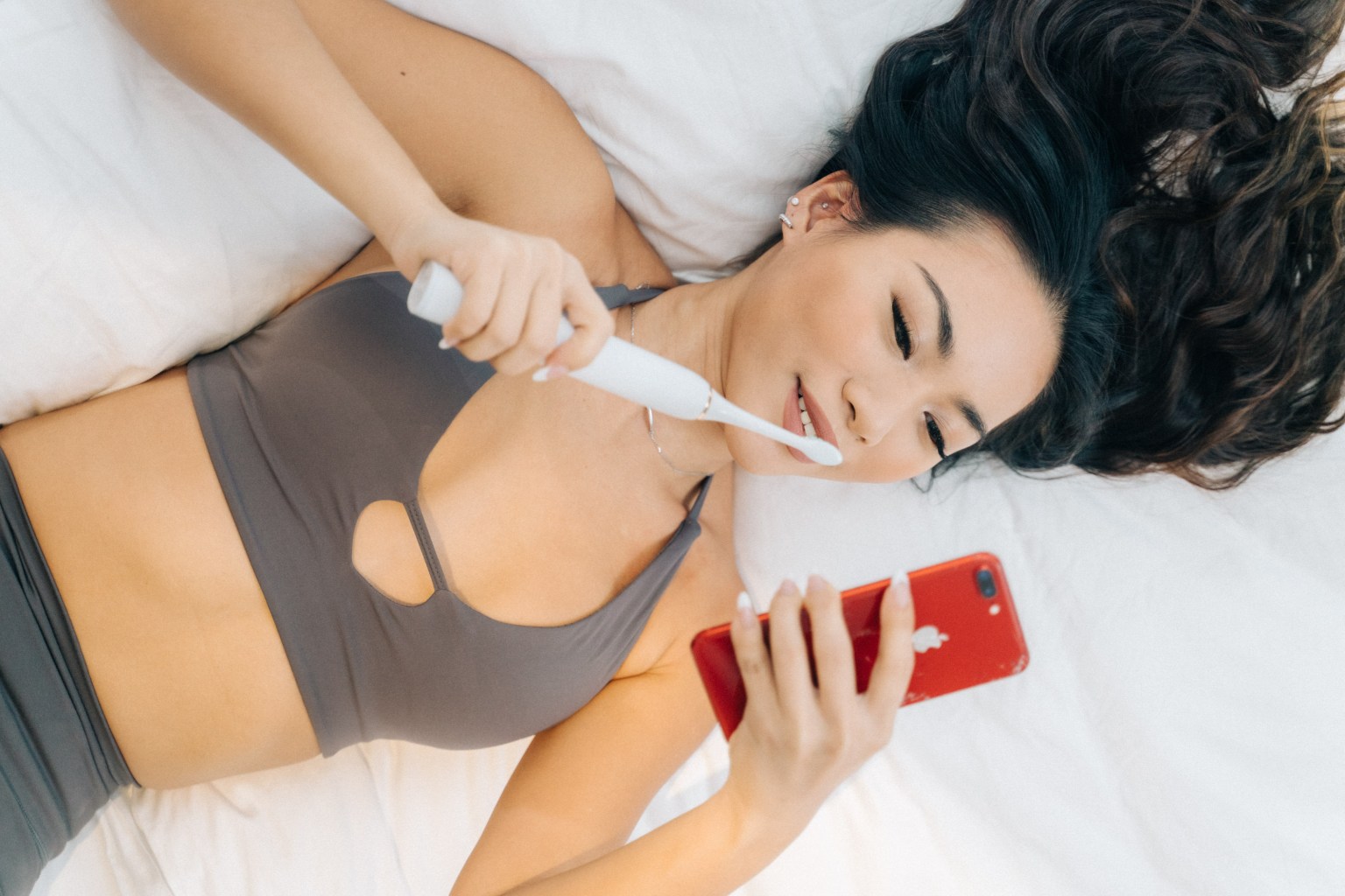 Young woman using an electric toothbrush while looking at her smartphone, lying on a bed, promoting dental care and patient engagement.