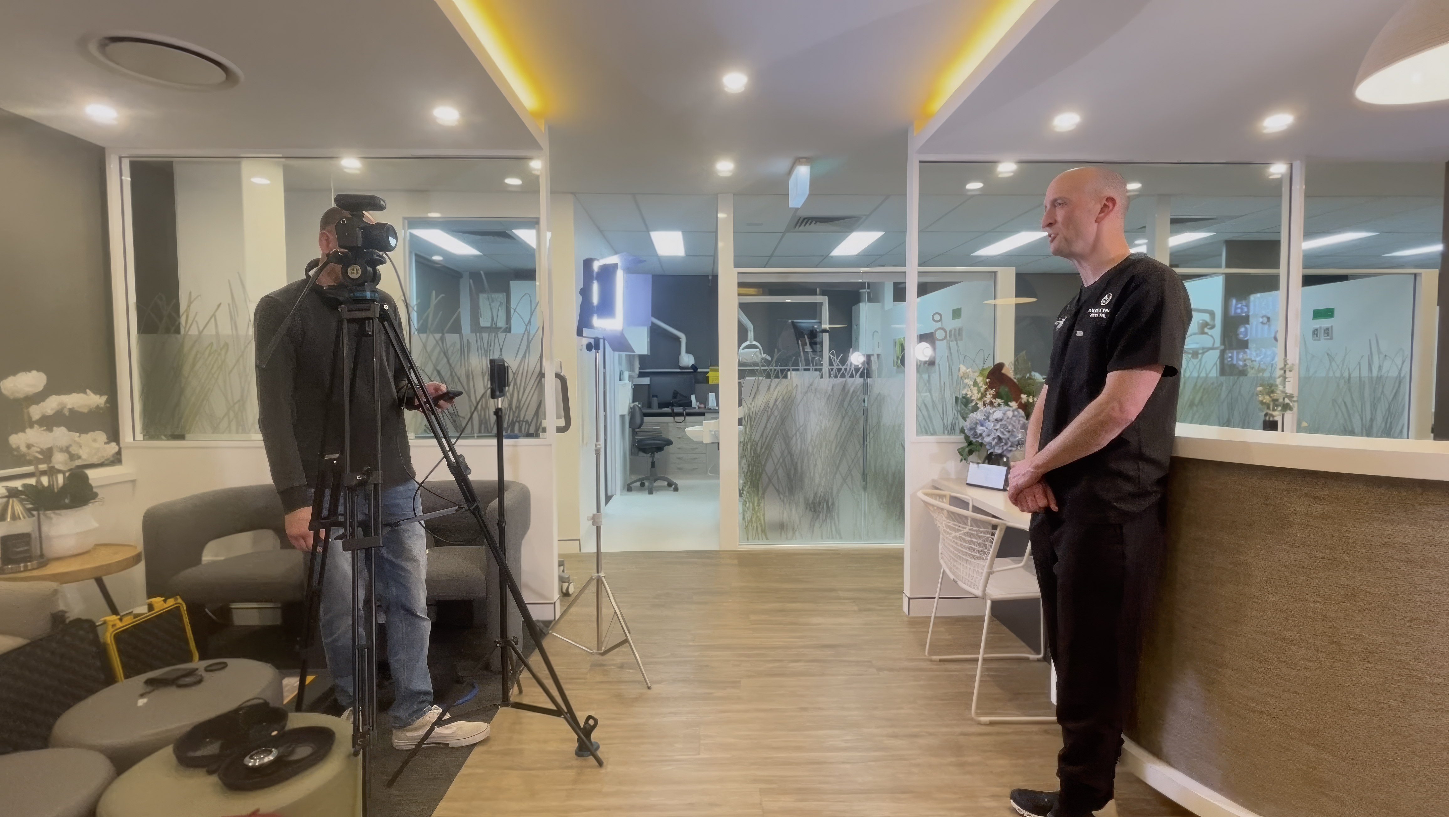 Dentist being filmed during a professional video shoot inside a modern dental practice, with camera, lighting setup, and clinical rooms visible in the background.